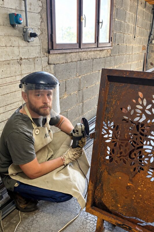 Artisan en tablier et visière de protection ponçant un panneau en acier découpé à motif de feuillage